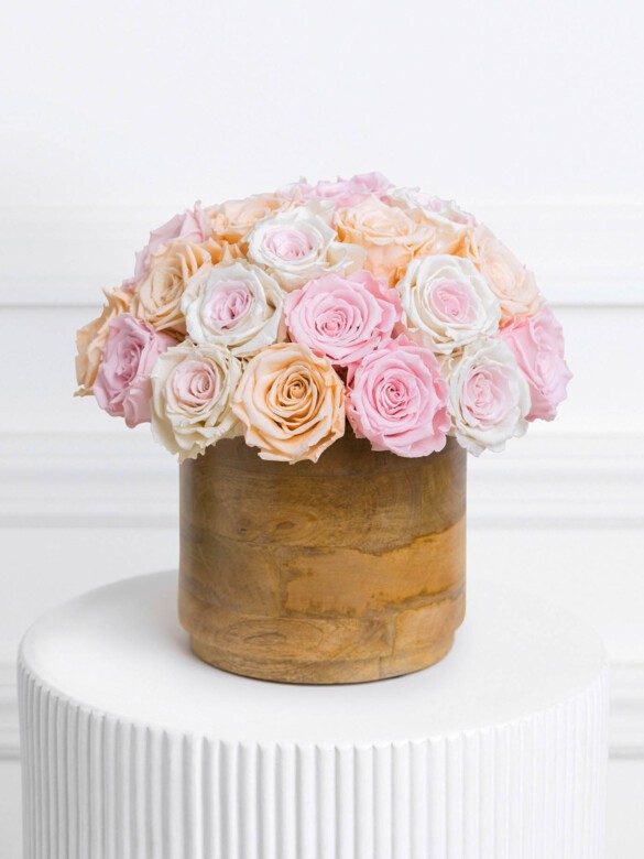 Pastel preserved rose arrangement featuring 30 soft pink, blush peach, and creamy white blooms, tightly clustered in a rounded silhouette atop a smooth wood-textured vase.