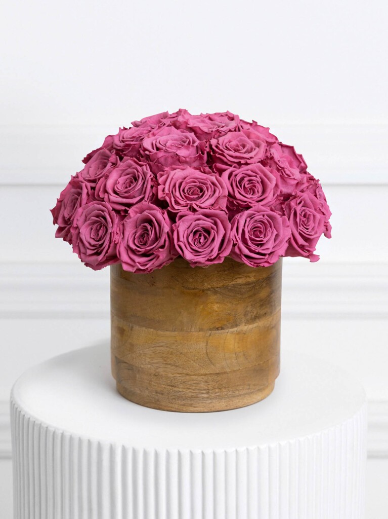 A lush dome of deep purple preserved roses blooms from a smooth, round wooden vase, each petal layered in crisp whorls for a sculptural, velvet-like effect against the natural woodgrain base.