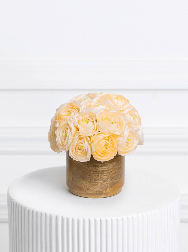 Compact arrangement of 21 pale peach preserved ranunculus, tightly clustered in a circular formation and presented in a smooth wooden cylinder vase, highlighting the flowers' ruffled texture and soft, sunlit glow.