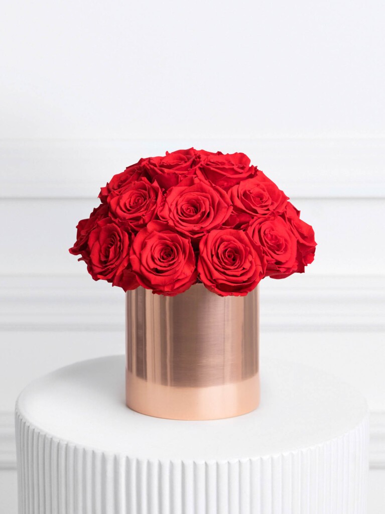Arrangement of vibrant red preserved roses tightly clustered in a tall, cylindrical metallic rose gold vase, forming a perfectly domed silhouette that blends modern luxury with timeless romance.
