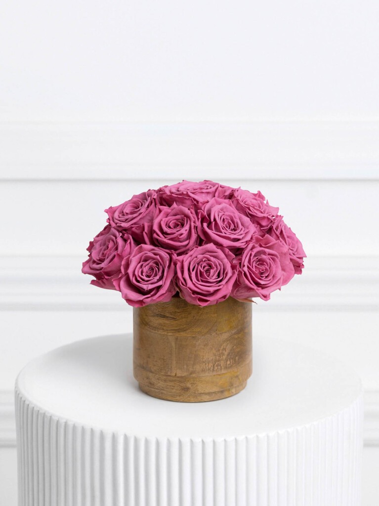 Vibrant arrangement of 16 preserved fuchsia roses displayed in a smooth, natural wood vase, with lush petals and evenly rounded shape creating a striking color contrast.