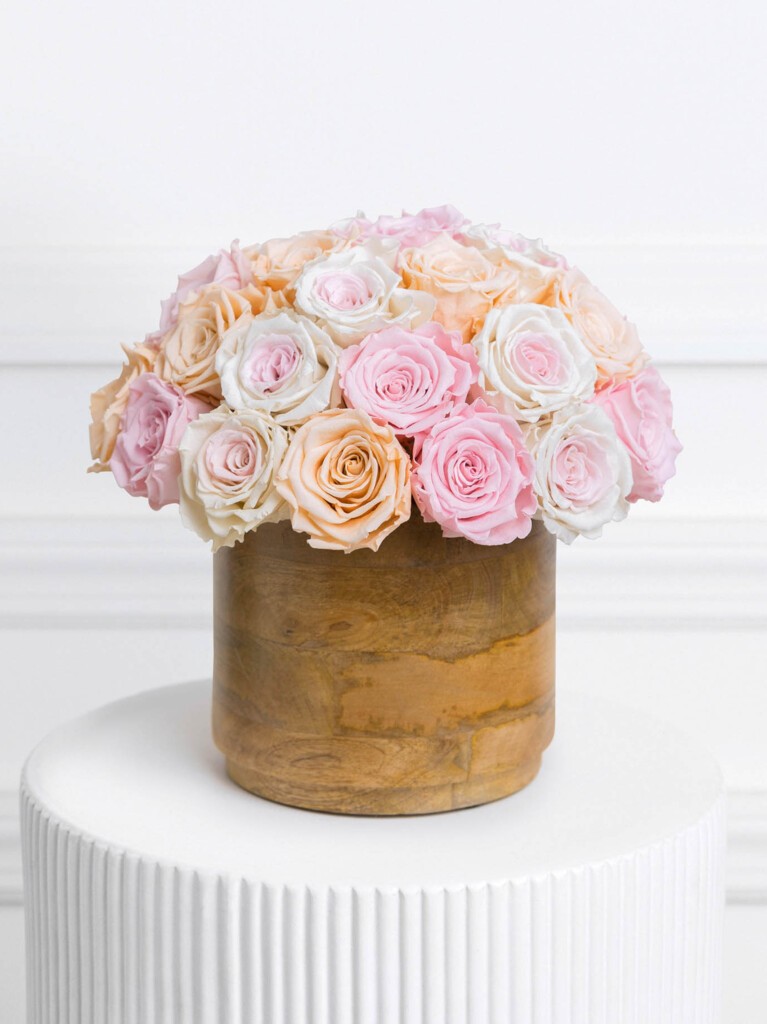 Pastel preserved rose arrangement featuring 30 soft pink, blush peach, and creamy white blooms, tightly clustered in a rounded silhouette atop a smooth wood-textured vase.