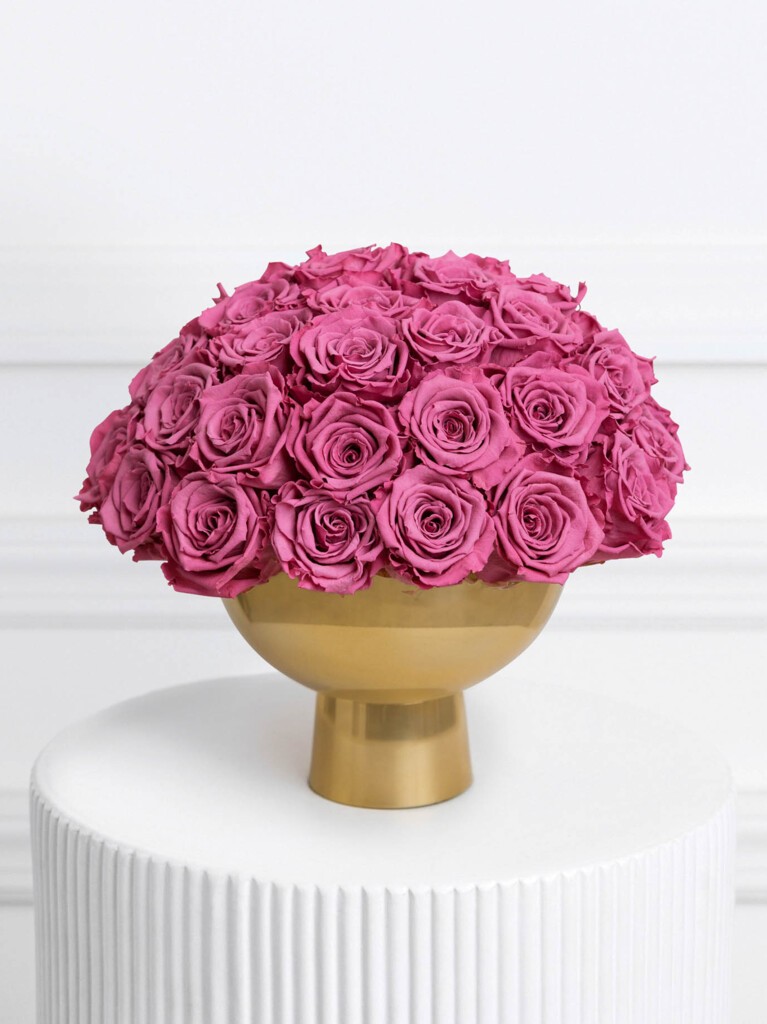 A lush dome of preserved roses in a deep raspberry pink hue, tightly arranged in a glossy gold pedestal vase.