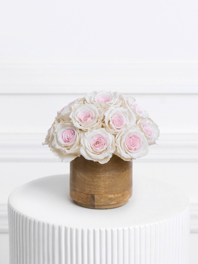 Preserved roses in a soft blush pink center with creamy white edges, arranged in a perfectly rounded dome and paired with a warm wooden vase for a romantic, two-tone composition full of texture and harmony.