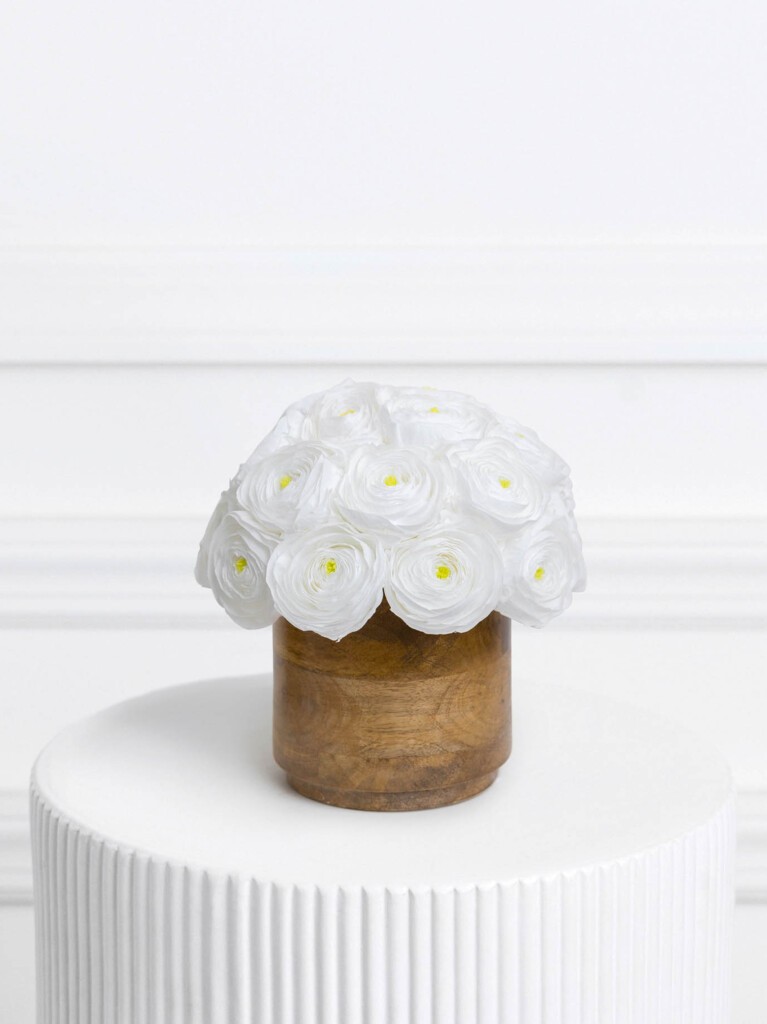 A compact dome of pristine white preserved ranunculus blooms with soft golden-yellow centers, arranged neatly in a smooth, natural wood-toned cylindrical vase for a warm, earthy contrast.