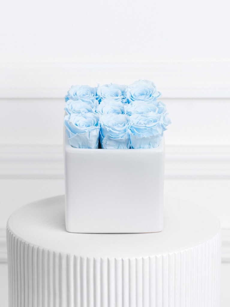A compact arrangement of soft baby blue preserved roses arranged in a tight 3x3 grid inside a smooth, modern white cube vase for a clean, dreamy, and minimalistic display.