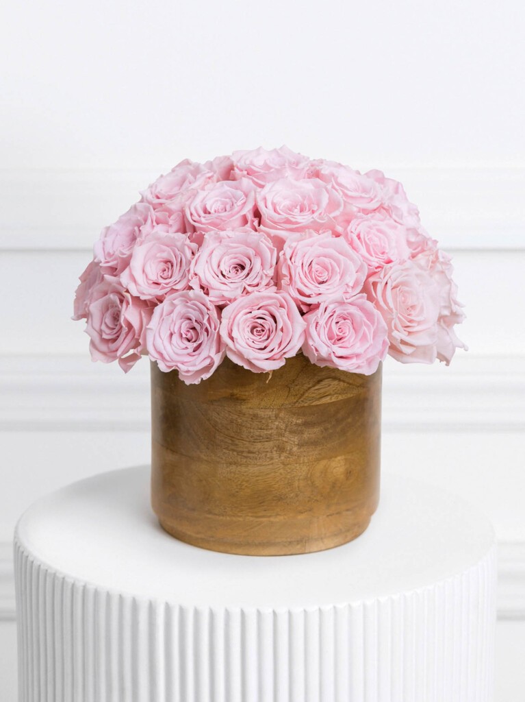 A lavish bundle of pale pink preserved roses blooming in a smooth, honey-toned wooden vase, combining timeless romance with a hint of rustic refinement.