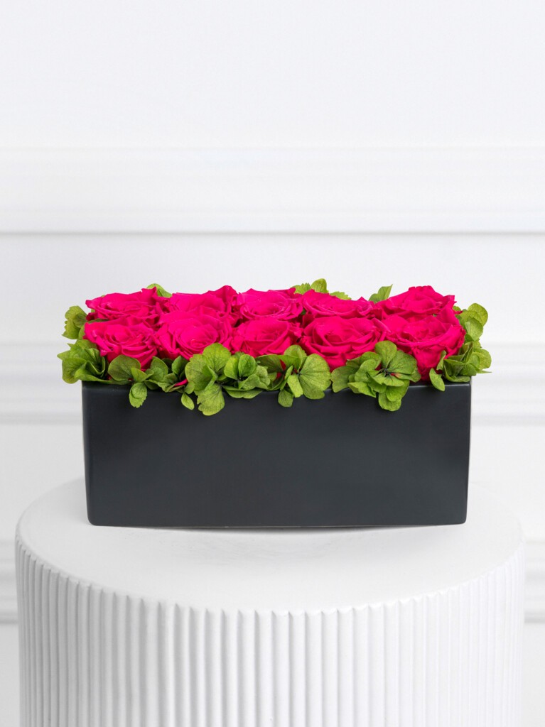 A low, rectangular arrangement of vibrant fuchsia preserved roses bordered by green hydrangeas, neatly styled in a modern matte black ceramic vase for a bold, contemporary look.