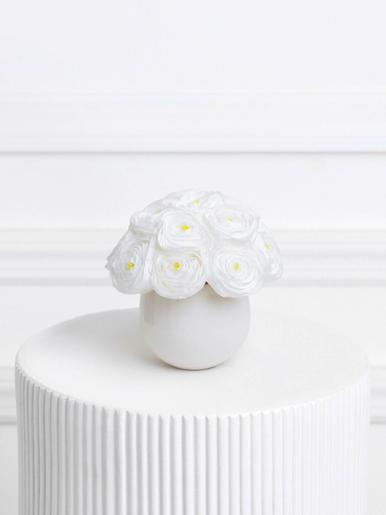 A compact bouquet of 14 preserved white ranunculus with soft yellow centers, arranged in a glossy white vase and displayed on a ribbed pedestal.